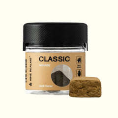 A small 3G clear jar of Nine Realms Classic La Mousse Hash with a black lid, shown next to a single brownish-green mousse cube on a light cream background.